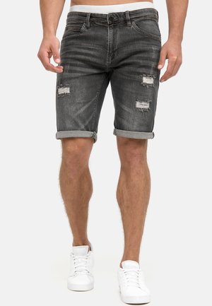 Homme portant un short en denim noir déchiré retroussé et des baskets blanches, debout devant un fond clair uni.