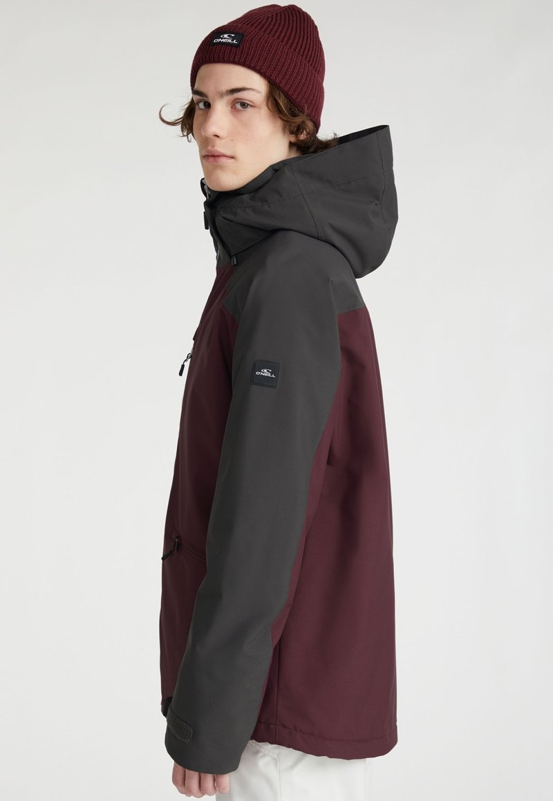 Maroon- und graue Jacke mit Kapuze, ausgestattet mit einer Reißverschlusstasche, verstellbaren Bündchen und einem Abzeichen am Ärmel. Texturiertes Gewebedetail.