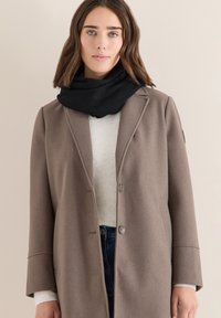 Abrigo de lana marrón claro con diseño entallado, bufanda negra y suéter de canalé crema. Cuenta con cierre de botones y bolsillos laterales.