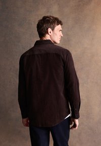 Ciemnobrązowa koszula z sztruksu z zapiętym na guziki kołnierzykiem, długimi rękawami i zaokrąglonym dołem. Charakteryzuje się gładką fakturą i subtelnymi detalami w postaci szwów.
