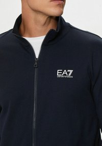 Uomo che indossa una giacca zip-up blu navy con il logo bianco EA7 Emporio Armani sul lato sinistro del petto sopra una maglietta bianca.