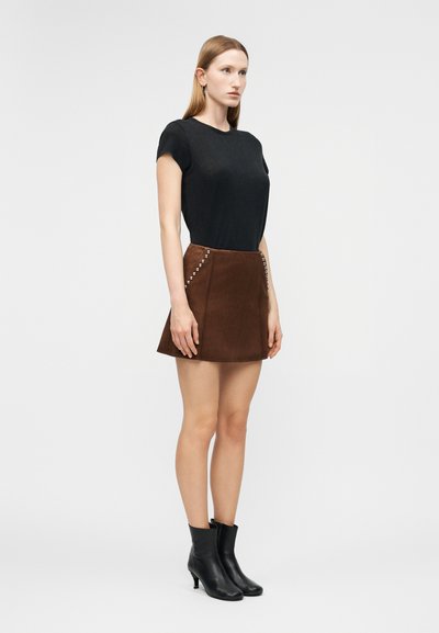 Γυναίκα που φοράει μαύρο κοντομάνικο τοπ, καφέ μίνι φούστα με τρουκς και μαύρα μποτάκια στον αστράγαλο, στέκεται μπροστά σε μια απλή φόντο.