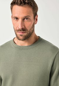 Uomo con capelli corti castani e barba, che indossa un maglione a girocollo di un verde chiaro e con texture, guarda diritto davanti a sé con un'espressione neutra.