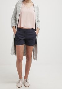 Světle růžový bezrukávový top, šedý texturovaný cardigan, námořnické šortky s vzorem a krémové oxfordky. Vyznačuje se volným střihem a vrstveným designem.