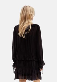 Vestido de gasa negro con mangas largas, capas con volantes y un detalle de lazo en los puños. Textura translúcida con un diseño suelto y fluido.