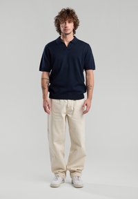 Tummansininen lyhythihainen polo-paita, jossa on V-kaula-aukko, yhdistettynä vaaleanbeigeihin leveälahkeisiin housuihin ja valkoisiin lenkkareihin, seisoo harmaata taustaa vasten.