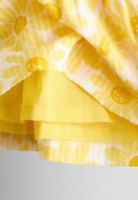 Material floral galben cu modele mari de margarete suprapuse peste material galben solid, prezentând cusături detaliate și textură.