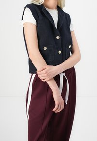 Tmav ě modrá pletená vesta se šálovým límcem, zdobená zlatými knoflíky a dvěma předními kapsami, doplněná vínovými kalhotami s bílými šňůrkami na stažení.