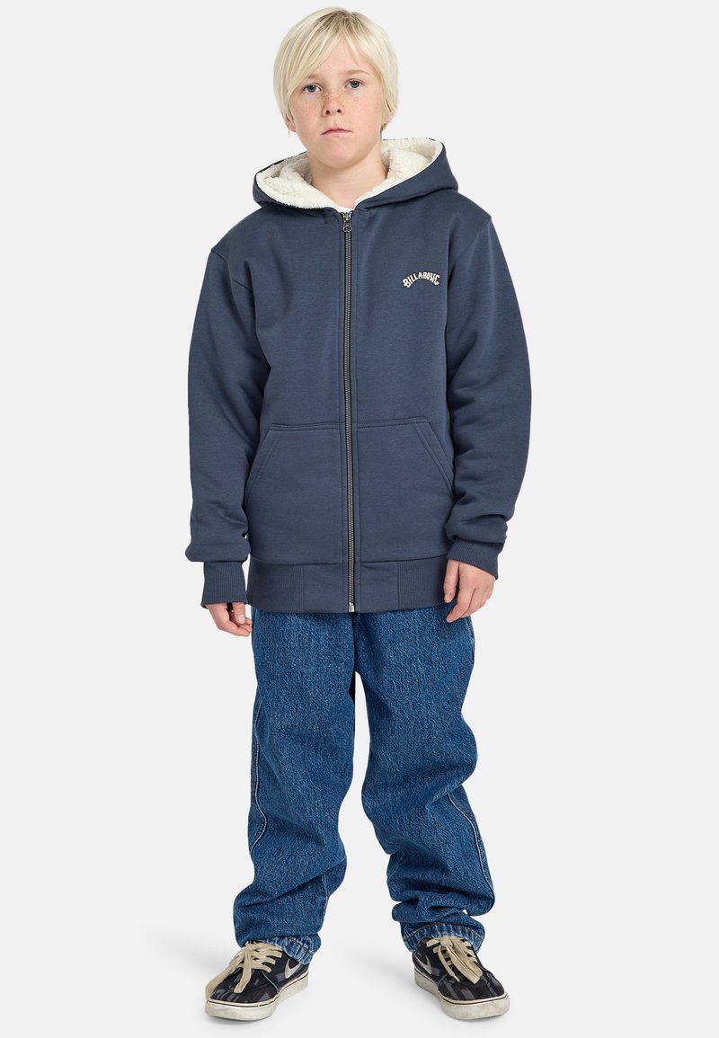 Felpa con zip di colore blu navy, foderata in pelliccia sintetica e con logo ricamato, abbinata a jeans in denim larghi e sneakers nere.