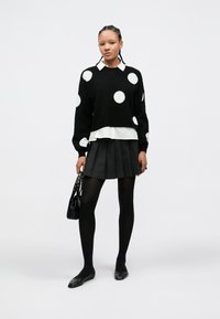 Camisola preta com bolinhas brancas de manga comprida, usada sobre uma camisa branca, combinada com uma saia preta plissada e collants pretos, complementada por uma mala.