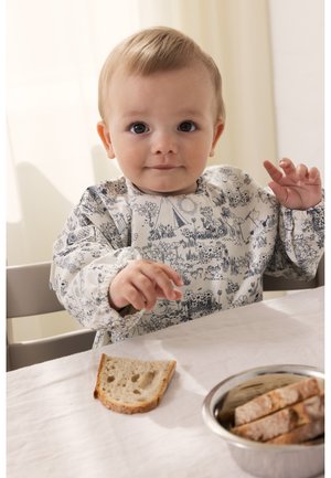 Malo dijete s uzorkom na portikla sjedi za stolom i pruža ruku prema kriški kruha, dok je u blizini zdjela s komadima kruha.