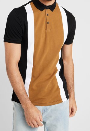 Mand iført en kortærmet polo med sorte ærmer og krave, brun front, hvide lodrette striber, kombineret med lyseblå jeans.