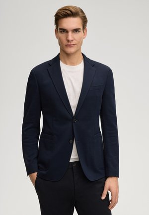 Giovane uomo con capelli castano chiaro che indossa un blazer blu navy sopra una camicia bianca, in piedi davanti a uno sfondo chiaro e uniforme.