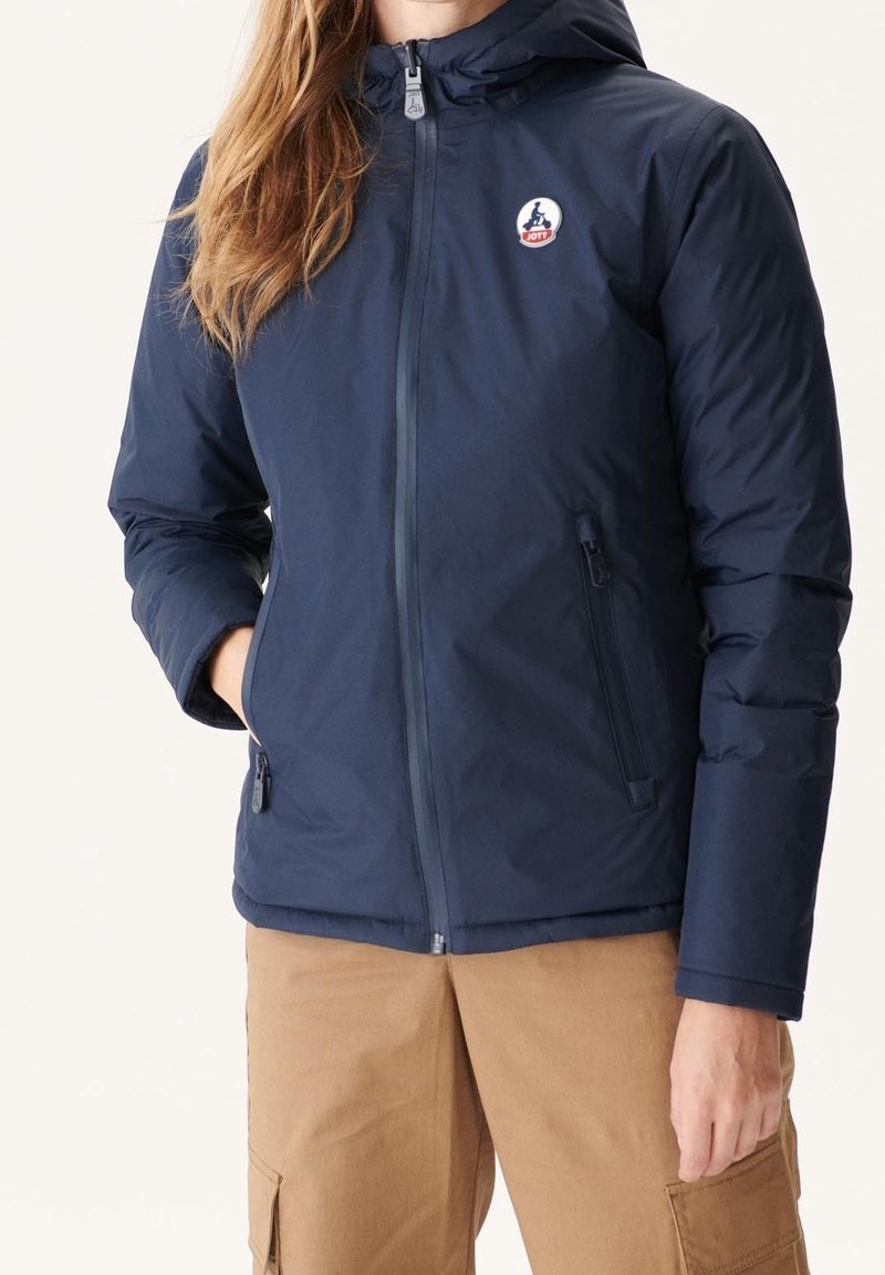Personne portant une veste à capuche bleu marine avec des poches zippées et un pantalon cargo beige, debout avec une main dans la poche.