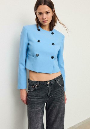 Junge Frau trägt ein kurzes, doppelt geknöpftes hellblaues Blazer und hoch taillierte, ausgewaschene schwarze Jeans, steht vor einer schlichten Wand.