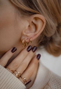 Pendientes de aro dorados en tres tamaños diferentes, mostrados contra un tono de piel claro; una mano con uñas pulidas de color oscuro descansa suavemente cerca de la oreja.