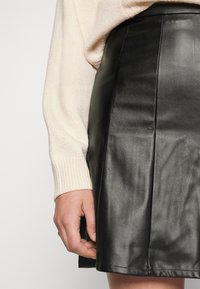 Svart läderkjol med en mjuk textur, med vertikala sömmar och en nederkant precis ovanför knäet, bärs tillsammans med en krämfärgad tröja.