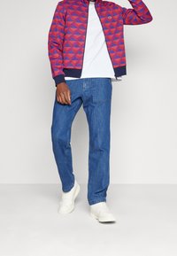 Veste zippée à motif géométrique rouge et violet, t-shirt blanc en dessous, pantalon en denim bleu, et baskets blanches. Style décontracté classique.