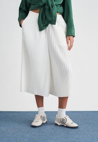 Personne portant un pantalon culotte large plissé blanc, un chemisier vert noué, des chaussettes blanches et des baskets blanches, debout sur un sol moquetté bleu.