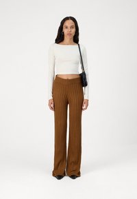 Gaiši bežs garām piedurknēm crop tops un brūni riboti plataji cimdi. Modelis tur nelielu melnu rokassomiņu. Gluda auduma un pieguļoša dizaina.