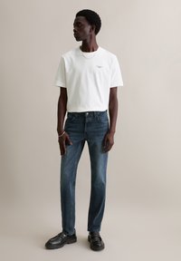 T-shirt blanc en coton, manches courtes, col rond ; jean en denim foncé avec une coupe droite ; chaussures en cuir noir avec une légère plateforme.
