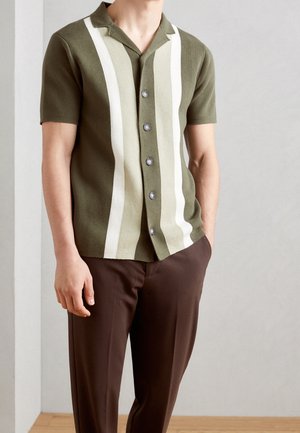 Mężczyzna ubrany w oliwkowozieloną, krótko rękawową koszulę na guziki z pionowymi białymi i jasnozielonymi paskami, w połączeniu z ciemnobrązowymi spodniami.