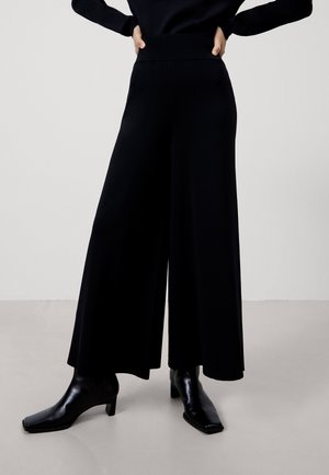 Persona con pantalones negros de pierna ancha, top negro de manga larga y botas negras brillantes hasta el tobillo, de pie con las manos en las caderas.