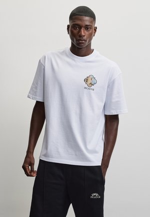 Mand iført hvid t-shirt med bjerglogo og teksten "PEGADOR", kombineret med sorte shorts med hvidt "PEGADOR" logo.