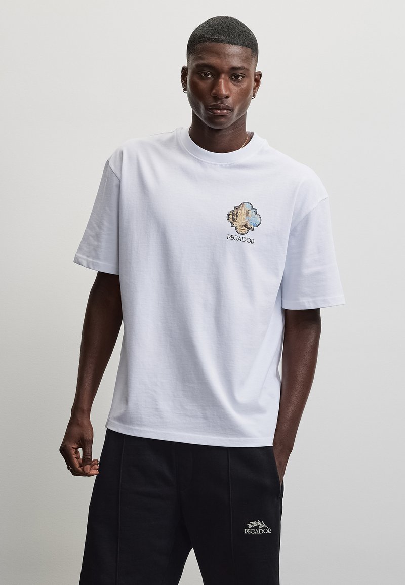 Homme portant un t-shirt blanc avec un logo de montagne et le texte "PEGADOR", associé à un short noir présentant le logo blanc "PEGADOR".
