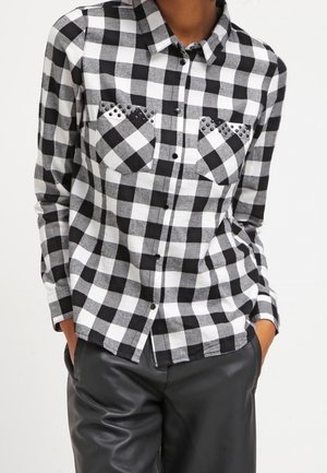 Zwart-wit geblokt overhemd met knopen en gestikte details op de zak, gedragen met zwarte leren broek, handen in de zakken.