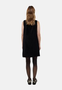 Robe noire sans manches avec une texture douce et lisse, présentant une coupe droite. Portée avec des chaussures à motifs et des collants noirs opaques.