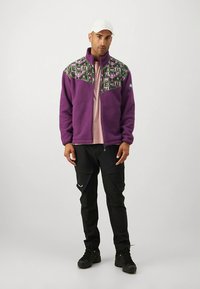 Vijolična jopa iz flisa z vzorčastim zeleno-črnim ovratnikom; kombinirana s črnimi cargo hlačami in škornji. Jopa ima visok ovratnik in zadrgo.