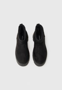 Une paire de bottes noires en suédine à chevilles avec des semelles en caoutchouc épaisses et des languettes de tirage, présentée vue de dessus sur un fond blanc.