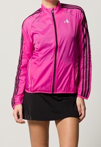 Chaqueta atlética ligera de color rosa con acentos negros, cierre delantero con cremallera, cuello alto y tres rayas negras en cada manga.