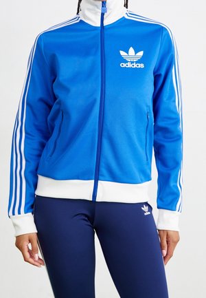 Personne portant une veste de survêtement Adidas bleue avec des bandes blanches et un logo, associée à un legging Adidas bleu marine avec un petit logo blanc.