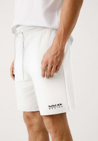 Shorts de algodón blancos con cintura elástica y cordón. Presenta un logo impreso de Red Bull Racing en la pierna izquierda. Textura suave.