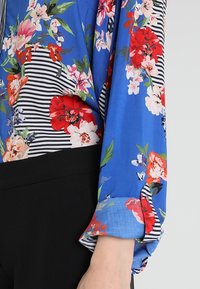 Blusa blu con motivi floreali rossi, rosa e arancioni e strisce bianche e nere, abbinata a pantaloni neri. Maniche arrotolate.