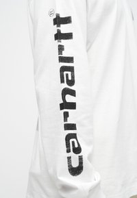 Hvid langærmet skjorte med fed sort tekst langs ærmet, lavet af bomuld og med en afslappet pasform. Teksten har et slidt udseende.