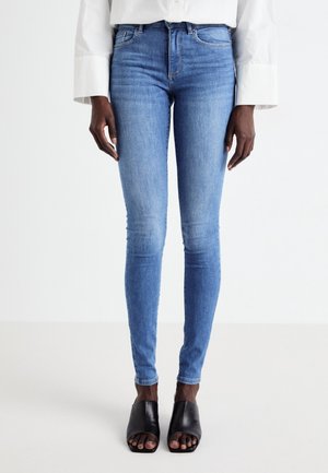 Femme portant un jean skinny bleu clair, une chemise blanche à manches longues et des chaussures à talons ouvertes noires, debout devant un fond blanc uni.