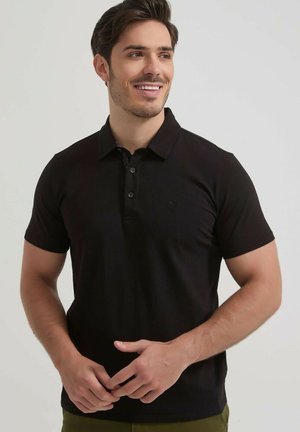Homme souriant portant un polo noir à manches courtes, debout les mains légèrement jointes devant un fond clair uni.