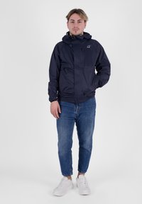 Giacca a vento navy con cappuccio, chiusura con zip e polsini elastici, abbinata a jeans blu e sneaker bianche. Design semplice e funzionale.