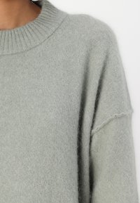 Hellgrüner Strickpullover mit geripptem Kragen, lockerer Passform und weicher Textur. Verfügt über gesenkte Schultern und subtile Stickdetails.