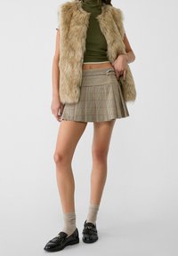 Beżowa kamizelka ze sztucznego futra na ciemnozielonym prążkowanym golfie, zestawiona z kratką mini spódnicą w beżowe, brązowe i czerwone tony; czarne mokasyny i skarpetki.