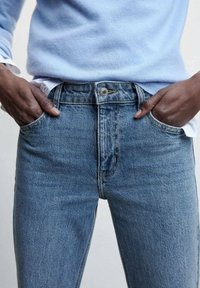 Jean en denim bleu à taille haute, design à cinq poches et fermeture à bouton. Le tissu a une texture légèrement usée et un aspect délavé.