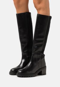 Bottes en cuir noir montant jusqu'au genou avec un panneau texturé noir, une fermeture éclair latérale, une semelle en caoutchouc épaisse et un talon bloc. Finition lisse avec un design durable.
