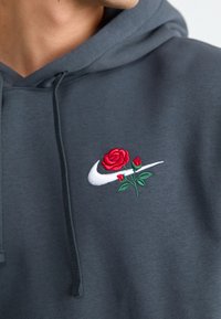 Grå hoodie med en vit Nike-swoosh och en röd ros samt gröna blad broderade på bröstet. Tyget är mjukt med dragsko.
