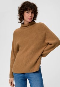 Brauner, gerippter Strickpullover mit hohem Kragen und überschnittenen Schultern, kombiniert mit blauen Jeans. Weiche Textur, lockere Passform und minimales Design.