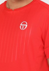 Punainen lyhythihainen urheilupaita, jossa pyöreä pääntie ja hienovaraiset pystysuorat raidat; ylävasemmalla valkoinen logo.