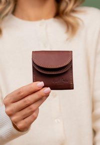 Roka, kas tur nelielu brūnu ādas monētu maciņu ar vāciņu, sīki izšūtiem šķautnēm un iedzīves zīmogu, cilvēks krēmkrāsas džemperī fonā.