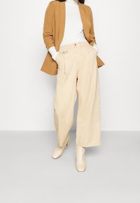 Pantaloni beige a gamba larga con pieghe frontali, abbinati a un dolcevita bianco e a un blazer marrone, completati da stivali con tacco a blocco beige.
